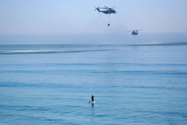 In this May 16, 2014 photo, a paddle boarder watches as two firefighting helicopters take on water from the Pacific Ocean off of San Clemente, Calif. The water-dropping helicopters were part of a ...
