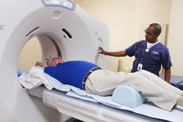 Mesfin Kidane, back, a nuclear medical technician, demonstrates how patient Brian Fong, 67, would enter the CT Optima 580 for a CT scan at Comprehensive Cancer Centers of Nevada Wednesday, Jan. 6, ...