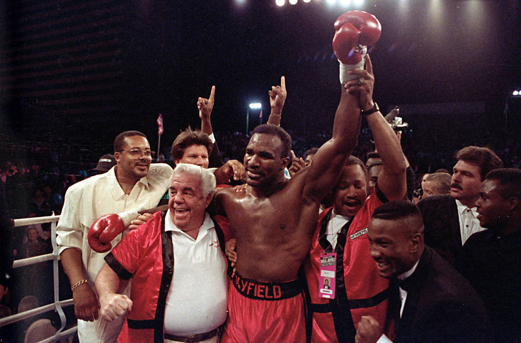 In this Oct. 25, 1990, file photo, the new world heavyweight champion Evander Holyfield is supported by his trainers Lou Duva, left, and George Benton, right, after his third round knock-out of Ja ...