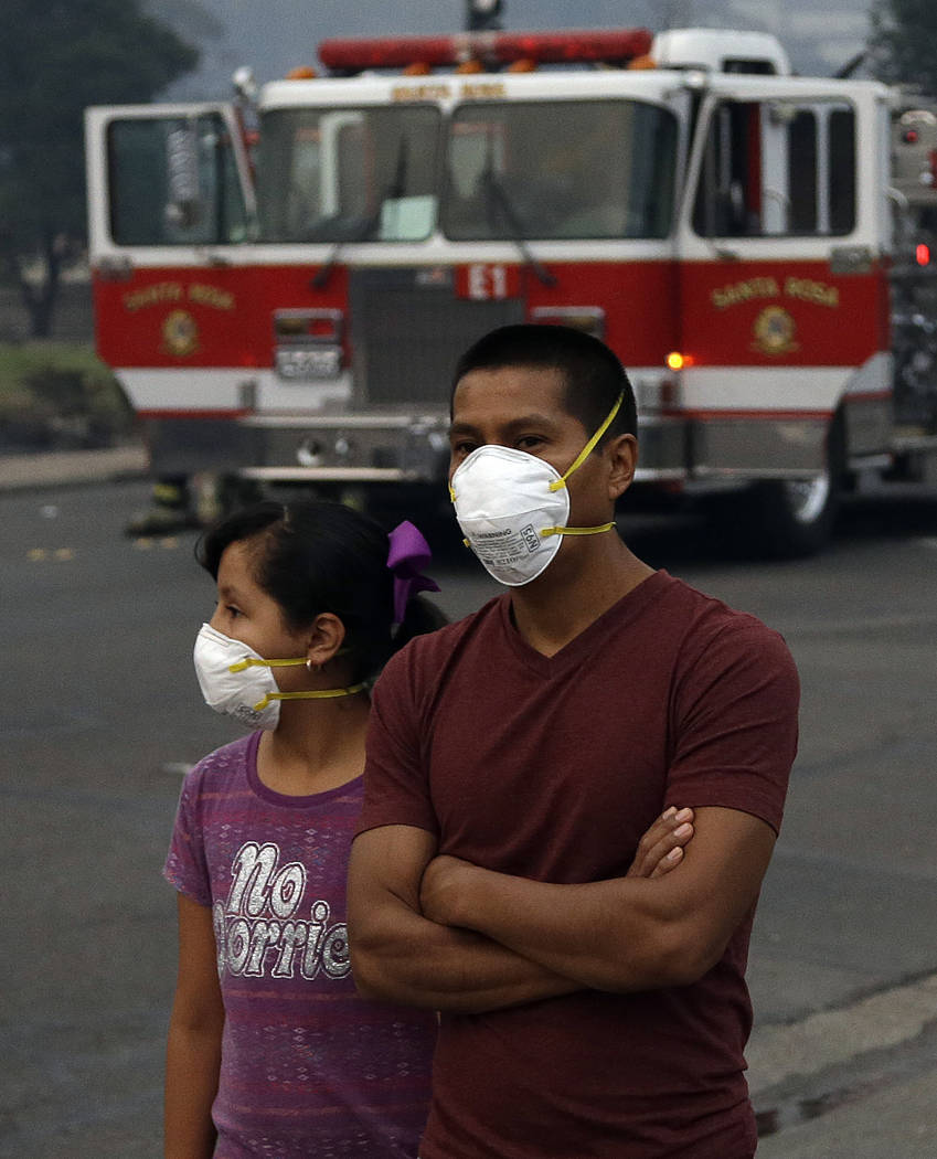 Pedestrians wear masks as they watch firemen battle a fire in a business on Monday, Oct. 9, 2017, in Santa Rosa, Calif. Wildfires whipped by powerful winds swept through Northern California early ...