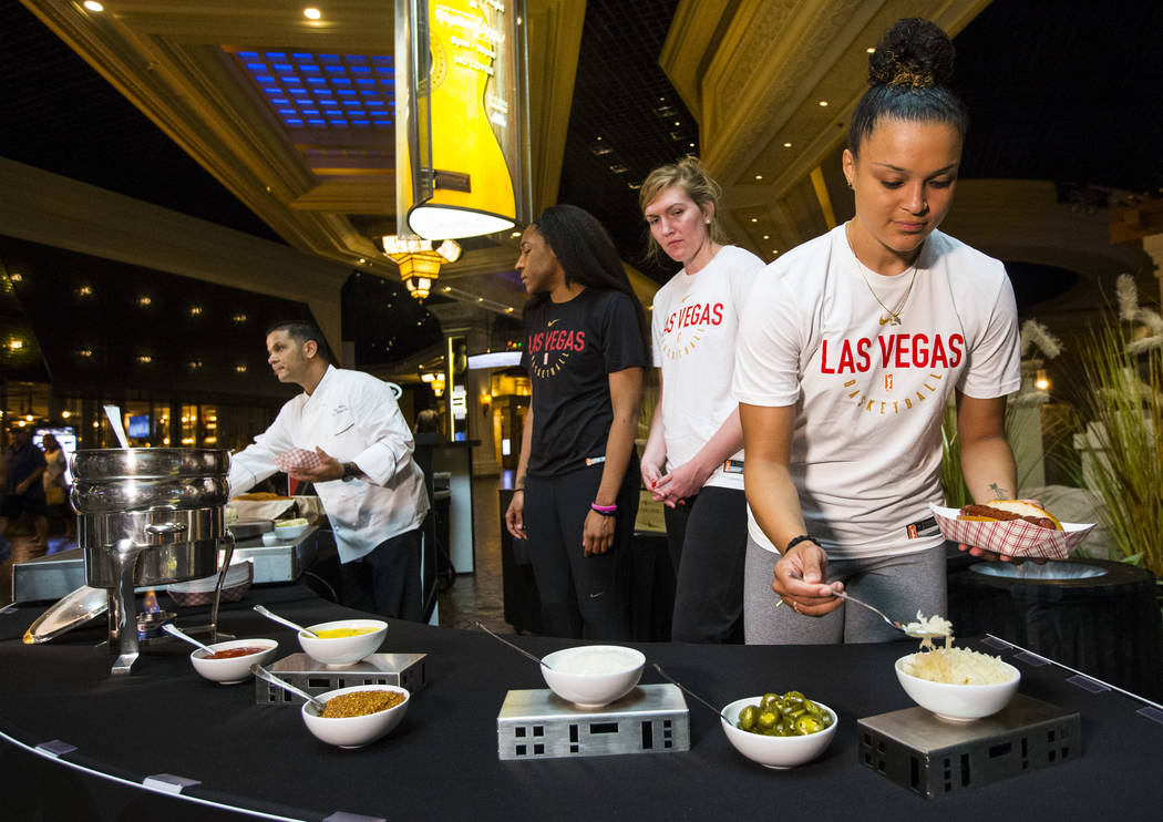 Las Vegas Aces player Kayla McBride, right, adds toppings to an "Aces Dog," a bacon-wrapped, mesquite-smoked beef dog in an herb garlic buttered bun with Jack Daniel's BBQ sauce, in the ...