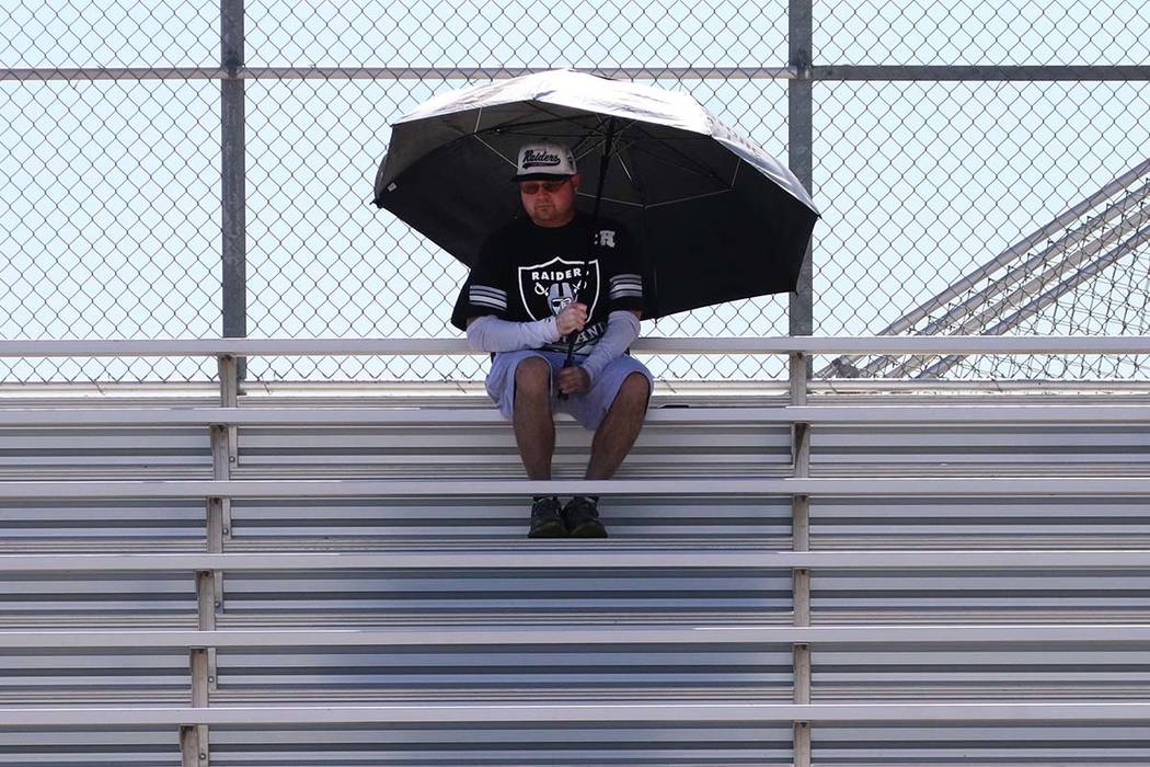 A man holds a giant umbrella to protect himself from sun as he watches football practice at Des ...