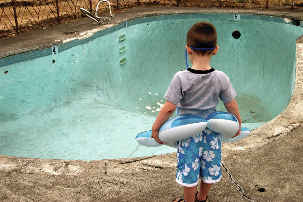 Small boy wearing a floaty and goggles stares at an empty swimming pool.
