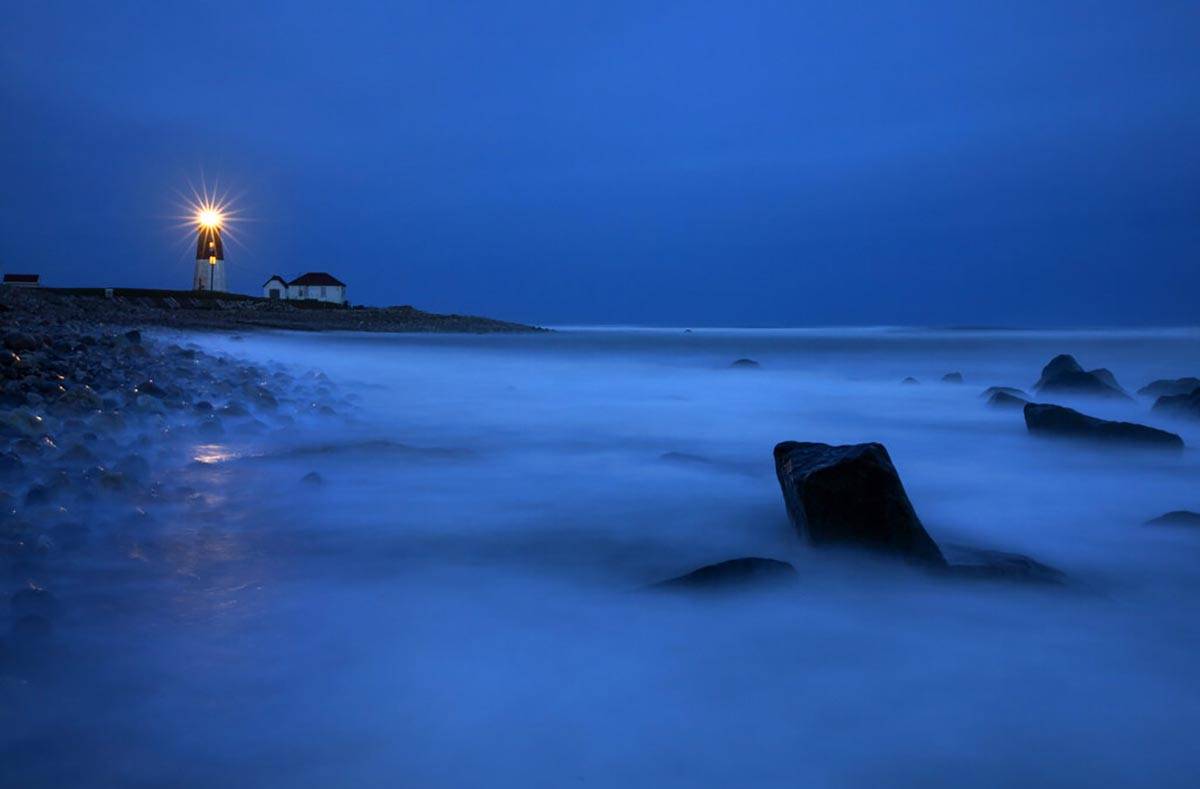 Waves wash over rocks as a beacon of light shines across the rocky shore from the Point Judith ...