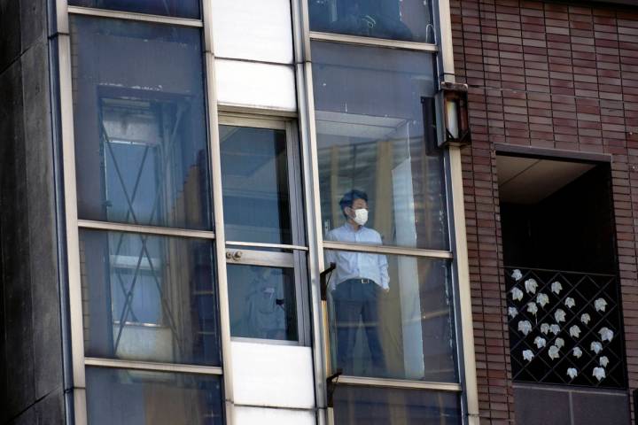 A man wearing a face mask to help curb the spread of the coronavirus stands at a building windo ...