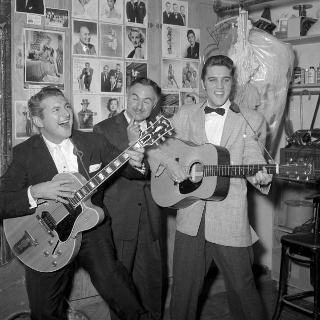 Liberace jokes around on a guitar as he and his brother George Liberace visit Elis Presley back ...