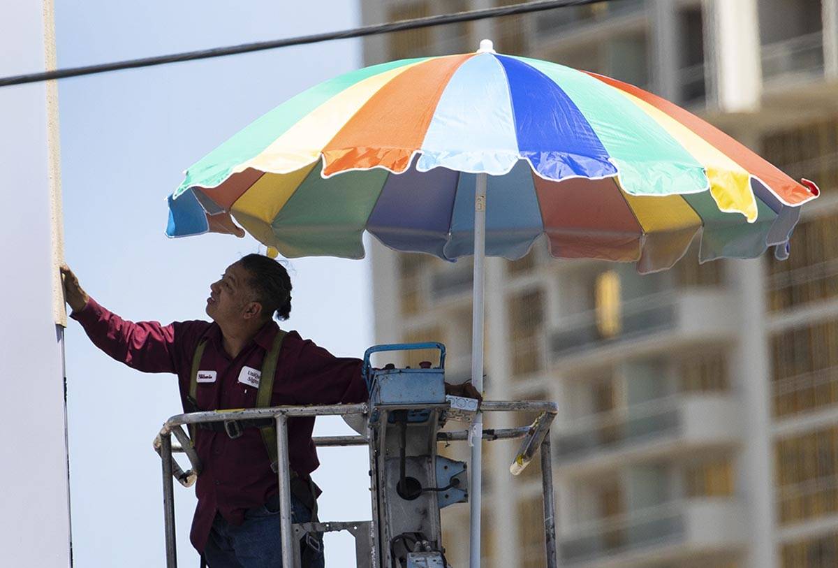 A worker uses a giant umbrella to protect himself from sun as he works on an outdoor advertisin ...