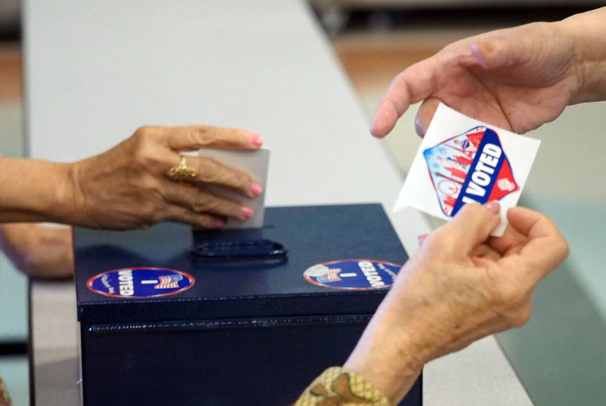A poll worker hands a voter a sticker showing that she has voted as another poll worker drops t ...