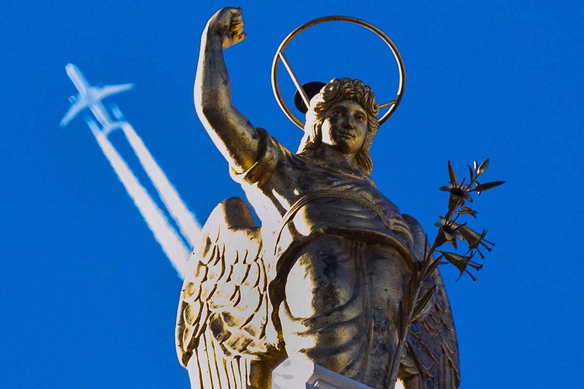 A passenger plane flies in clear blue sky and leaves a white trail as it flies over a statue in ...