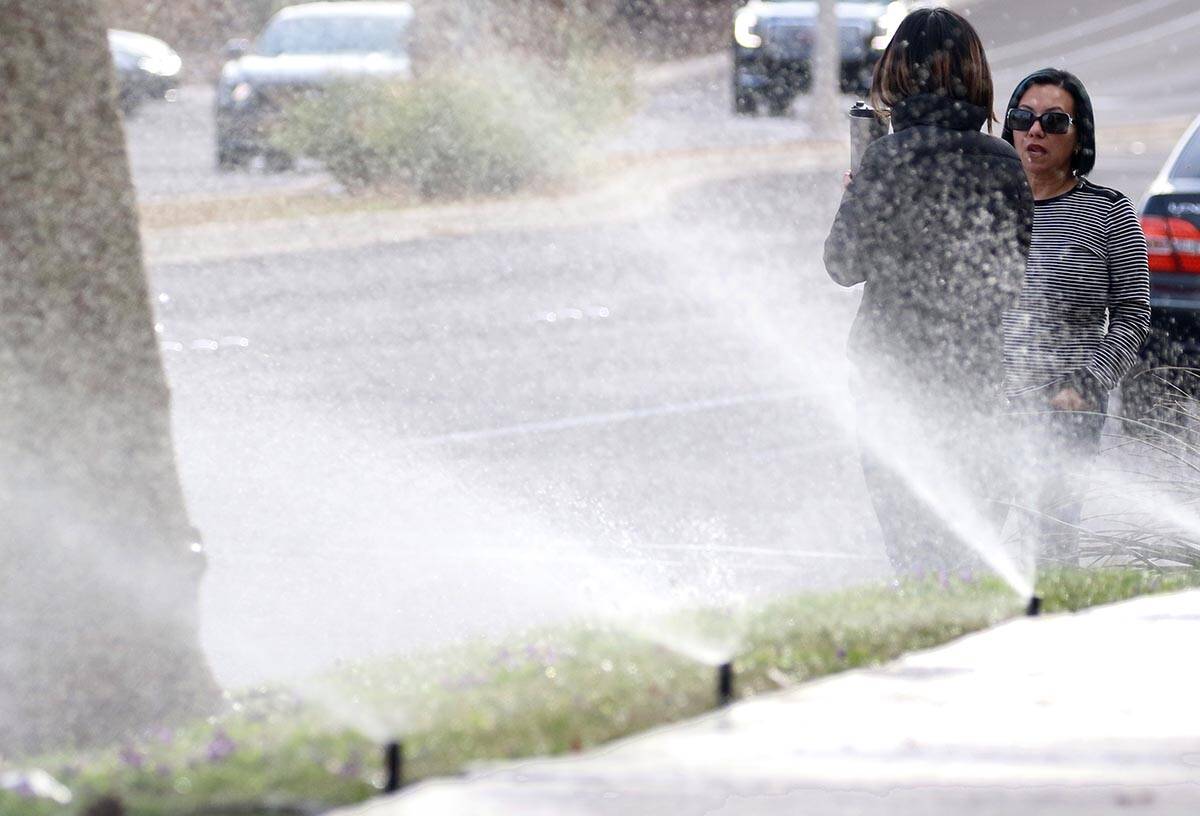 Pedestrians wait for green light to cross Green Valley Parkway as lawn sprinklers are on to wat ...