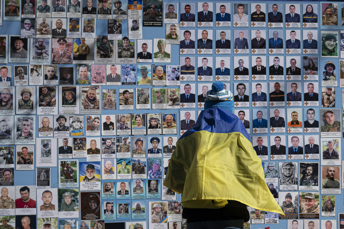 A woman wrapped in a Ukrainian flag stands next to the Memory Wall of Fallen Defenders of Ukrai ...