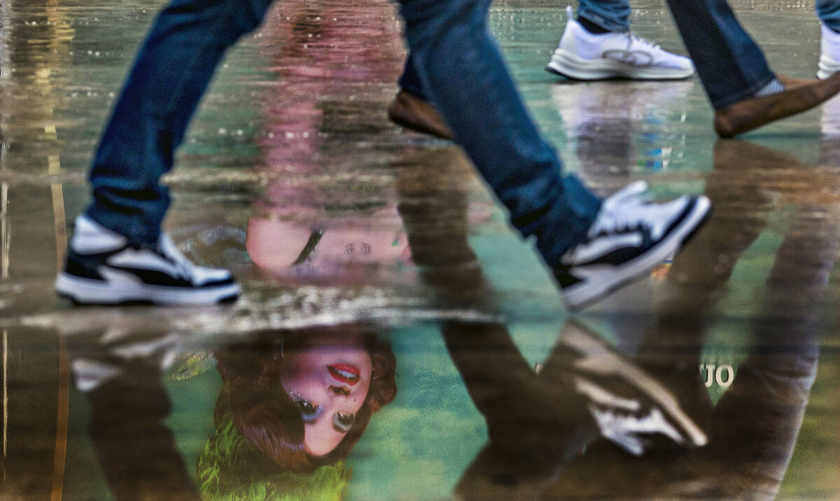 The Green Fairy from the show Absinthe is reflected in a puddle as pedestrians walk past the en ...