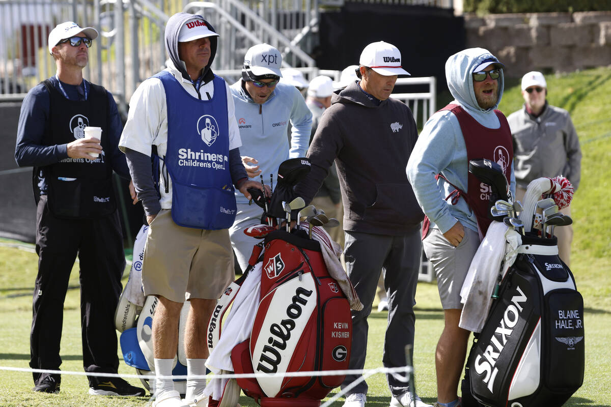 Players and their caddies are bundled up to protect themselves from heavy wind as they wait to ...