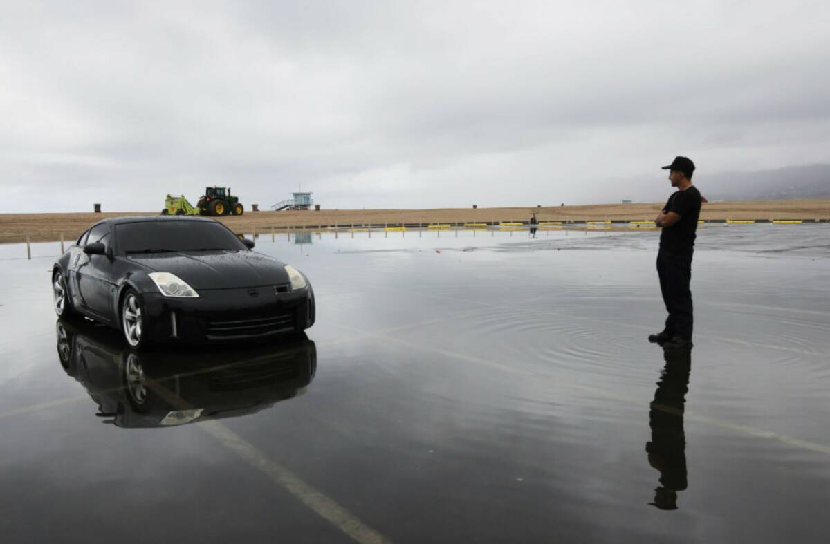 A man stands alongside a car in a flooded beachside parking lot after heavy rains Tuesday, Oct. ...
