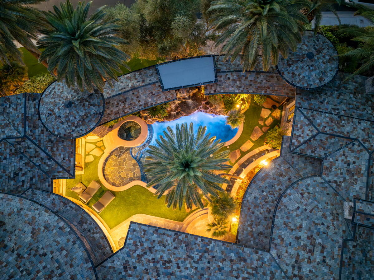 Overhead drone view of the lit pool, lawn, palm tree, and stone patio in the backyard of the MacDonald Highlands home at dusk