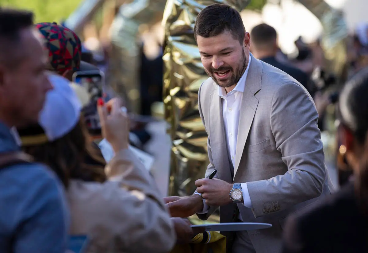 A smiling Golden Knights player in a light gray suit signs a puck for a fan on the gold carpet before the Golden Knights' 10th season opener